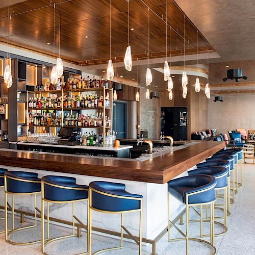 The image shows a modern bar with a sleek wooden countertop, stylish chairs, hanging lights, and a fully stocked liquor shelf in a spacious, well-lit setting.
