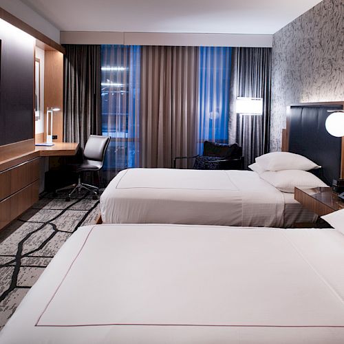 A modern hotel room with two double beds, a wall-mounted TV, a desk with a chair, and contemporary decor.