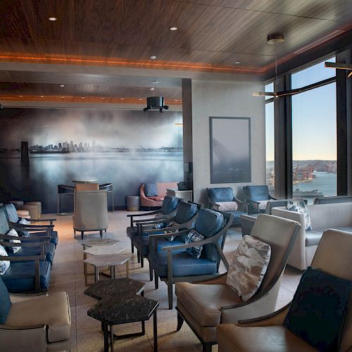 A modern lounge area with cushioned chairs, tables, large windows offering a scenic view, and a wall art piece.