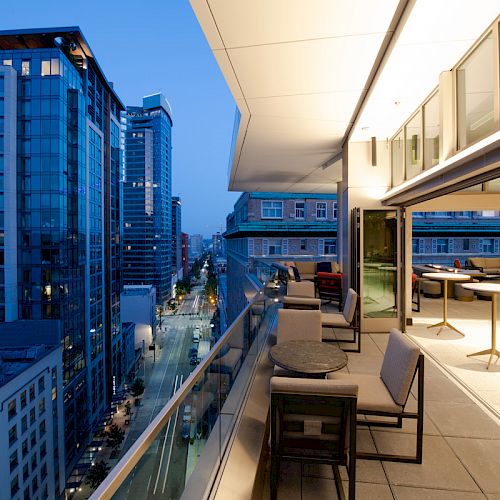 An urban balcony view at dusk shows modern city buildings, tables, and chairs on the terrace, with a partially open glass wall revealing indoor seating.