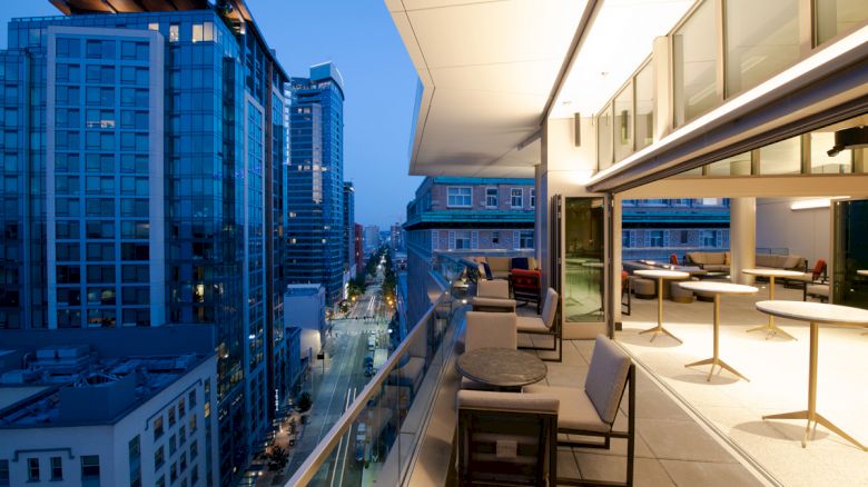 The image shows a modern cityscape in the evening with tall buildings and an illuminated, stylish rooftop terrace with seating and tables.