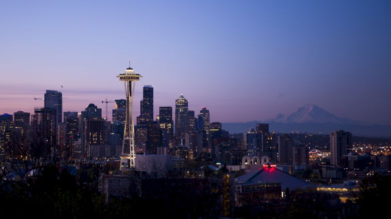 The image shows the Seattle skyline at dusk, featuring the Space Needle prominently with Mount Rainier in the background and city buildings.