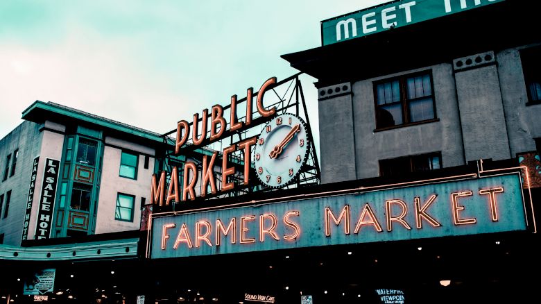 The image features a neon sign for a public market and farmers market in front of buildings, under a partly cloudy sky.