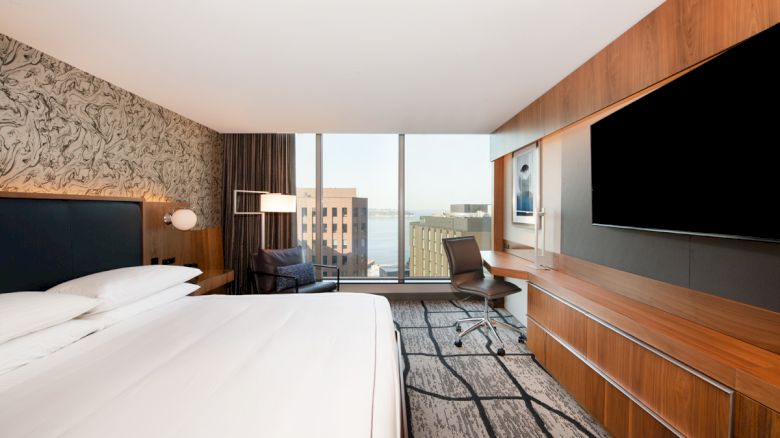 The image shows a modern hotel room with a large bed, a wall-mounted TV, a desk with a chair, and a window overlooking city buildings.