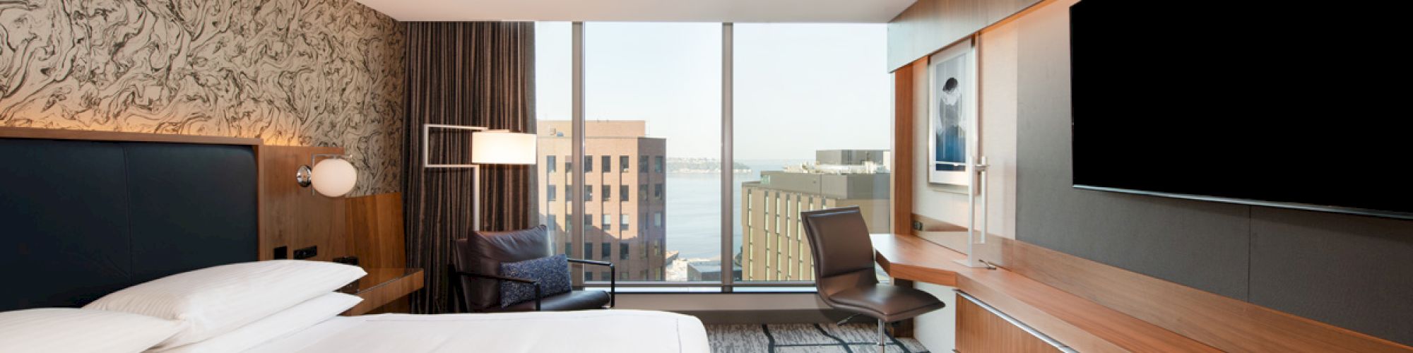 The image shows a modern hotel room with a large bed, a wall-mounted TV, a work desk with a chair, and a floor-to-ceiling window with a city view.